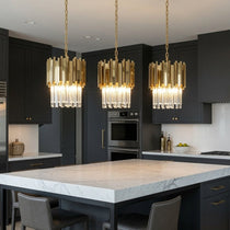 Three modern cylindrical pendant lights with gold-toned rods and clear glass shades hang over a white marble kitchen island in a contemporary dark gray kitchen.